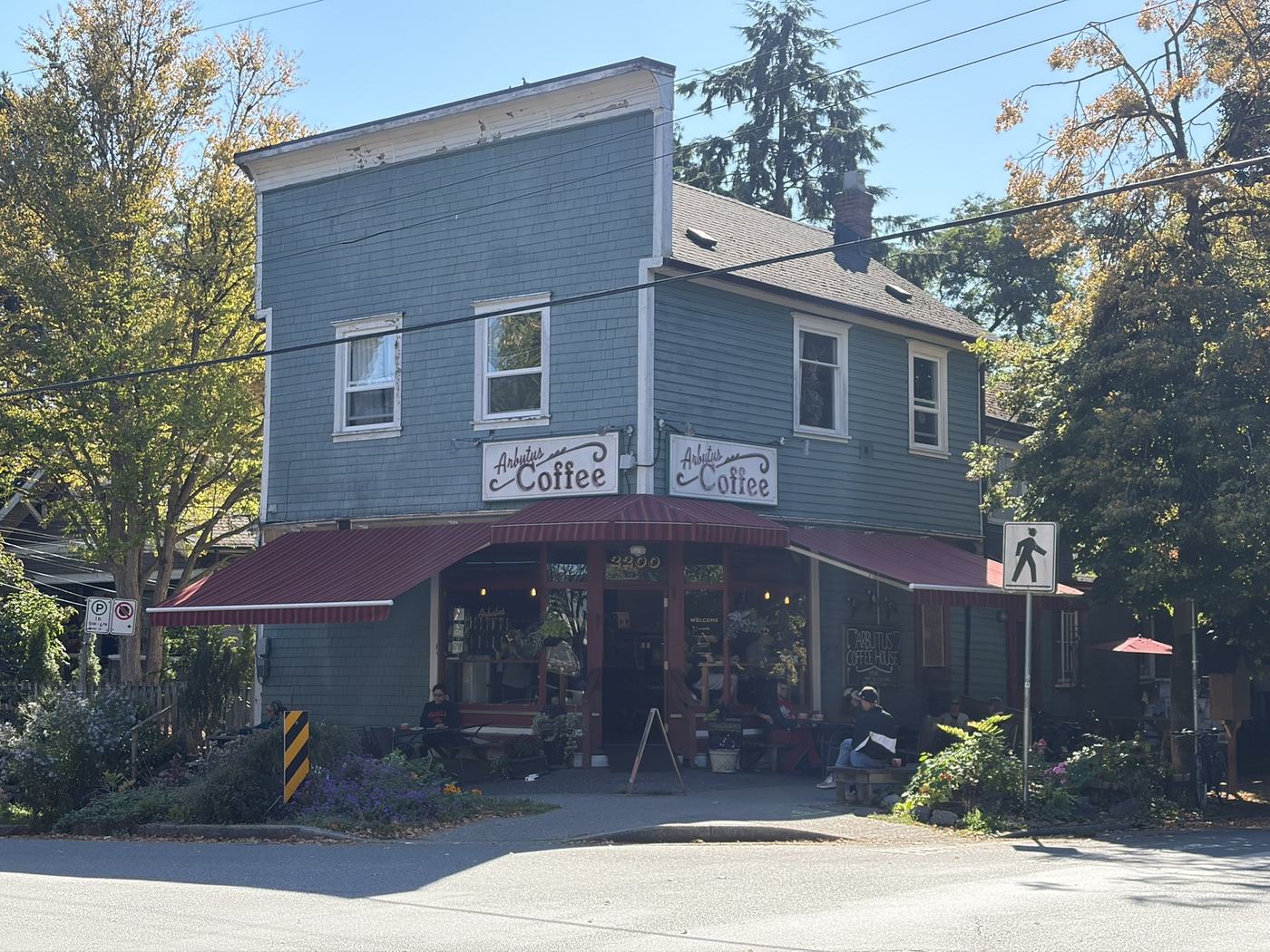 A two-storey blue-grey wooden corner building with two painted Arbutus Coffee signs above a dark red awning, and a few people sitting at tables outside
