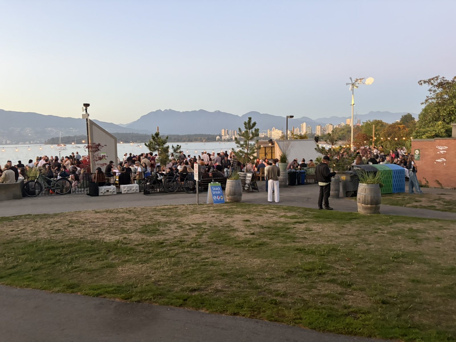A crowded outdoor patio beside Kitsilano Beach at dusk, with string lights, barrel planters and the North Shore mountains across the water
