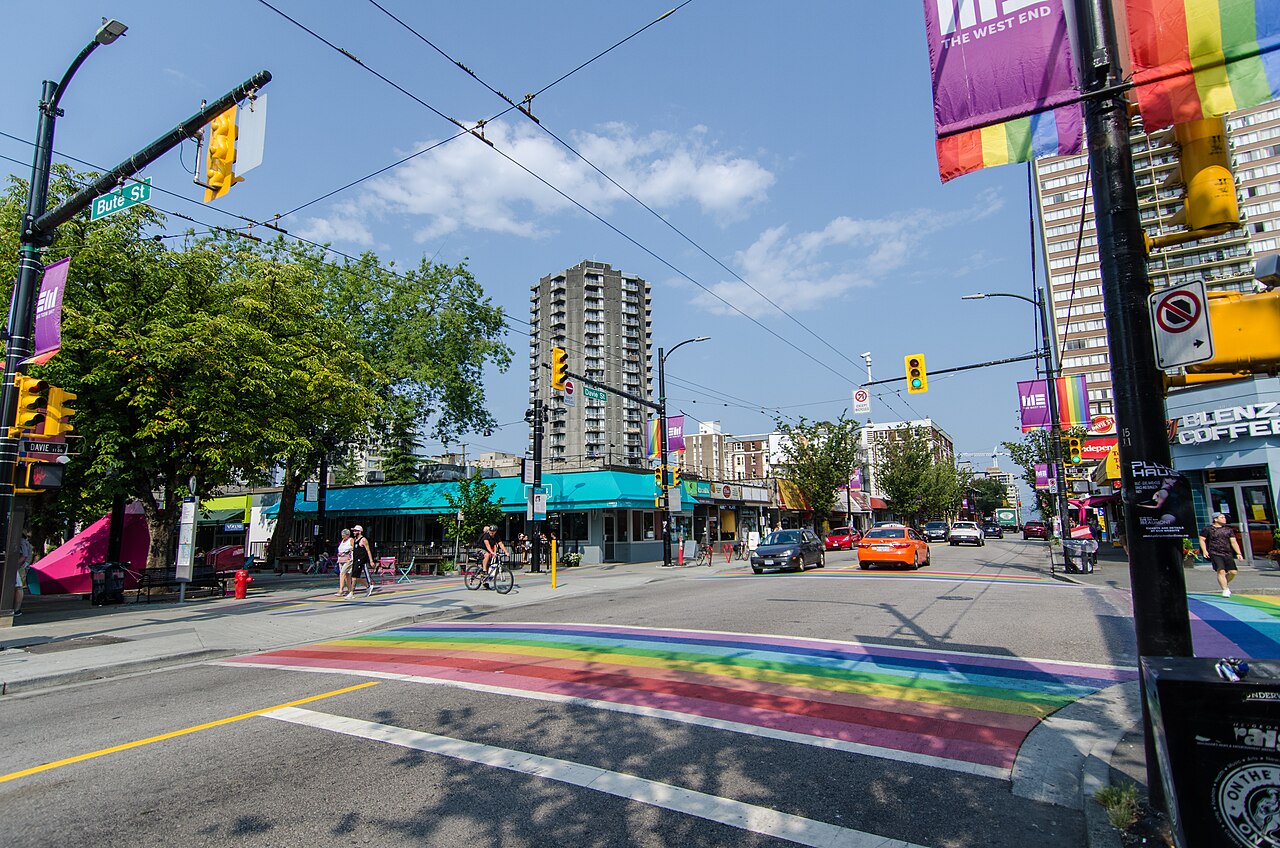 Davie Street at Bute Street, with rainbow crosswalks, sidewalks, shops and traffic on a sunny day