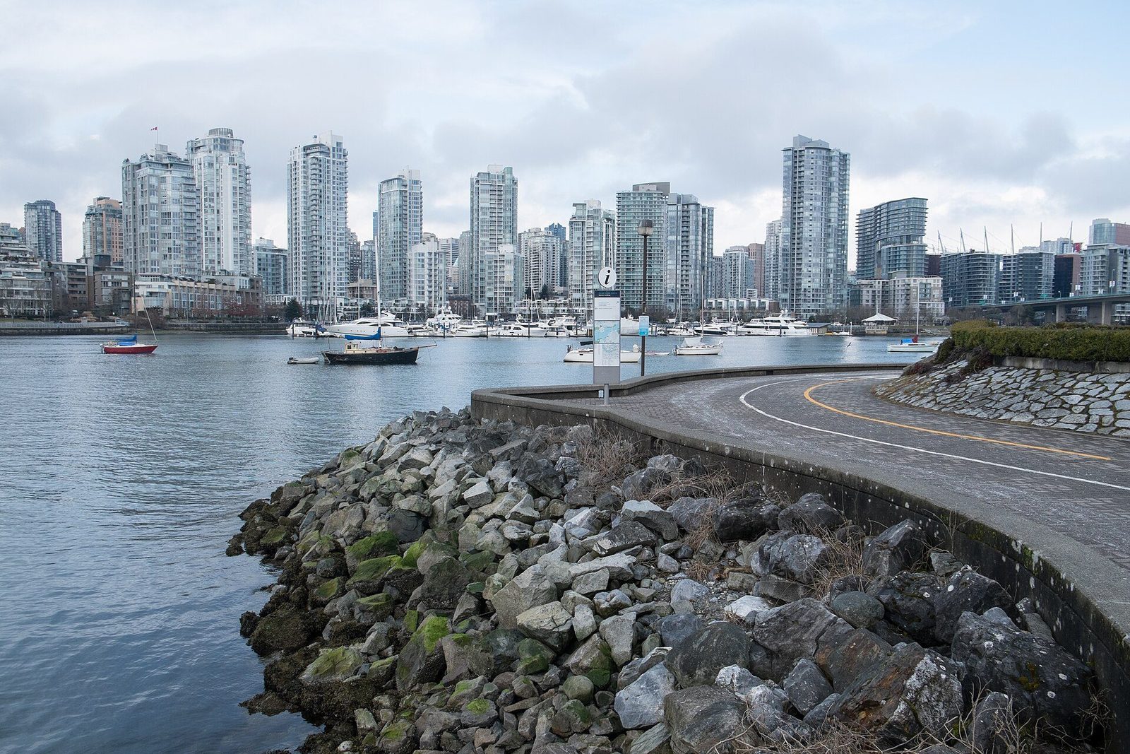 A curving seawall path along rocks at the edge of False Creek, with a skyline of downtown condo towers across the water
