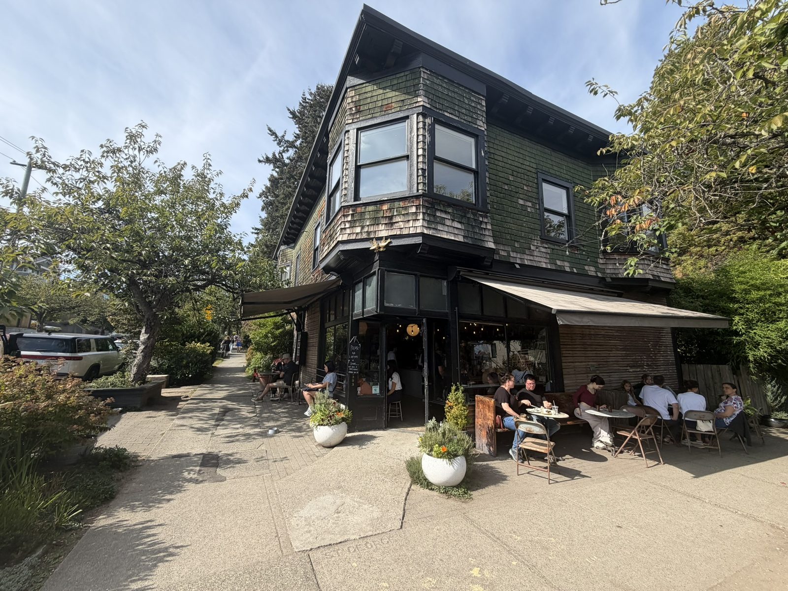 A two-storey shingled corner building with dark trim and bay windows, with people at patio tables under a canvas awning