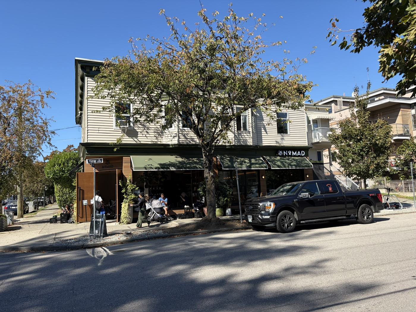 A café with dark green awnings on the ground floor of a cream two-storey building, with people sitting at the open frontage